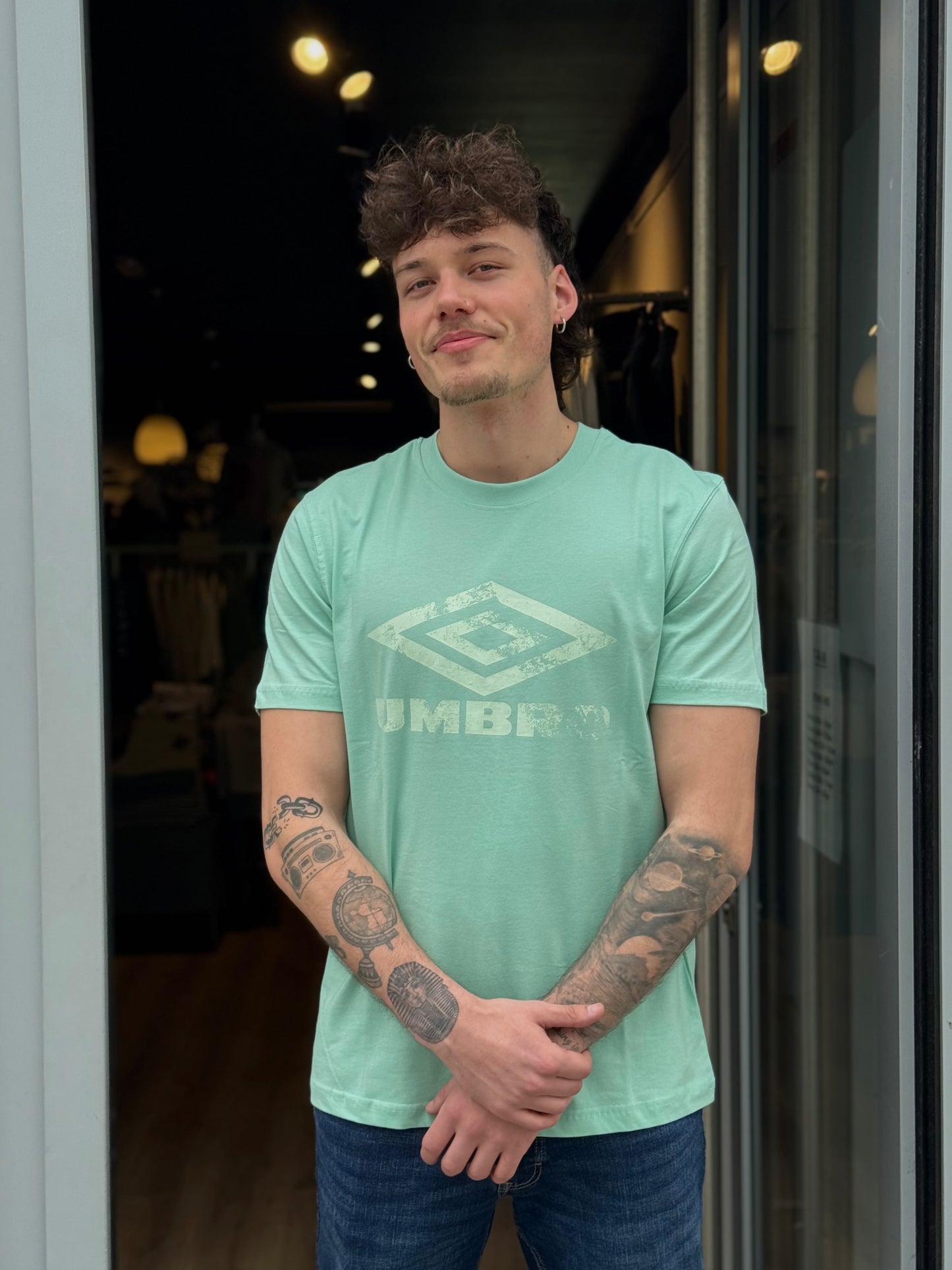 Man wearing a light green Umbro t-shirt standing in front of a glass door.