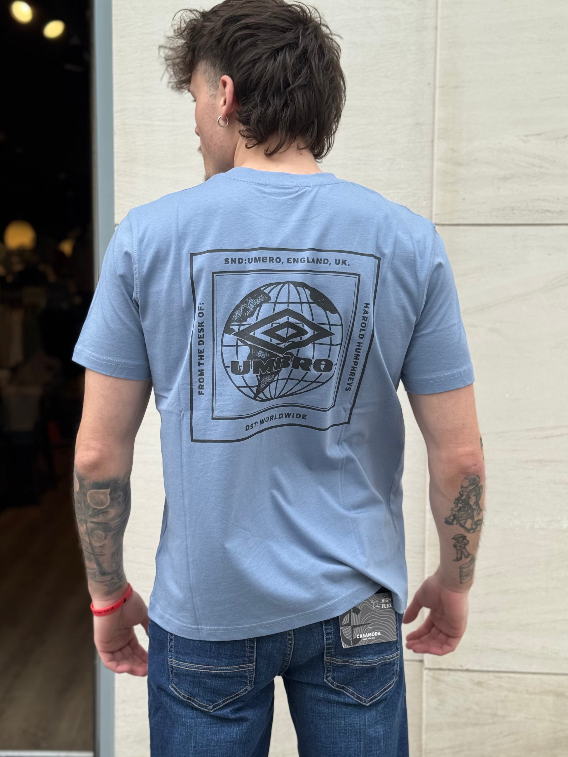 Man wearing a light blue t-shirt with a graphic design on the back, standing against a light-colored wall.