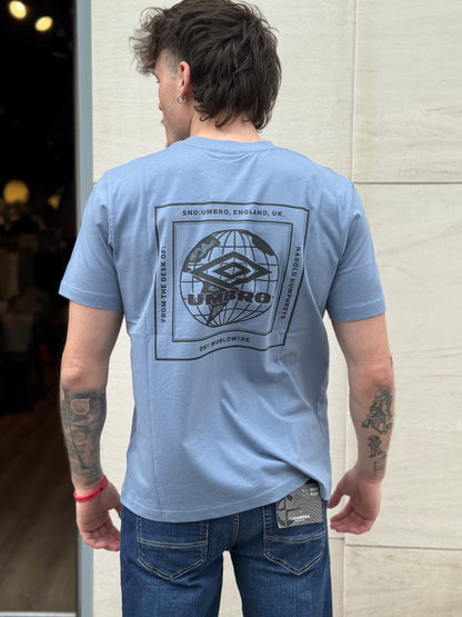 Man wearing a light blue t-shirt with a graphic design on the back, standing against a light-colored wall.