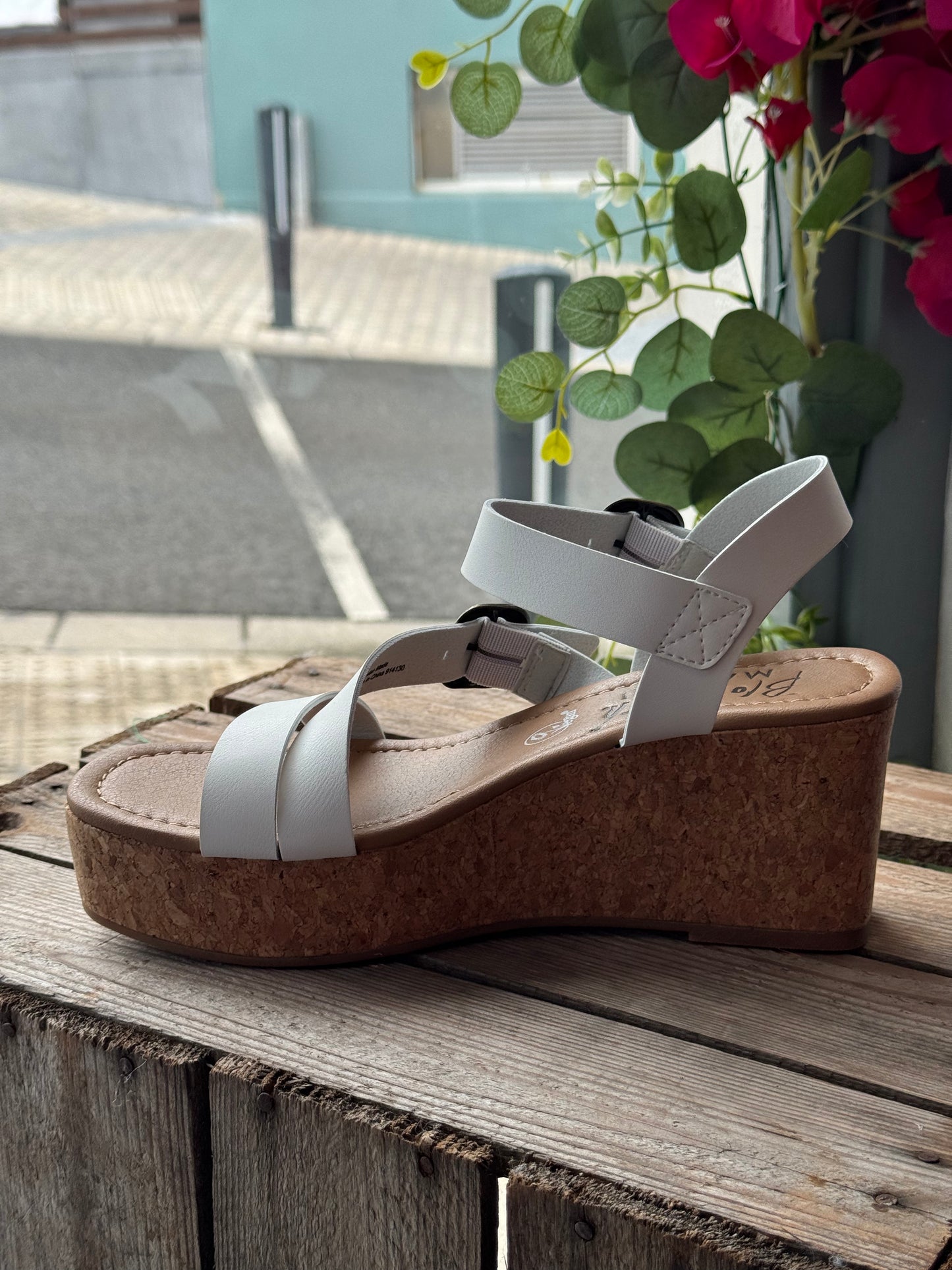Brown wedge sandal with white straps on a wooden surface, with plants in the background.