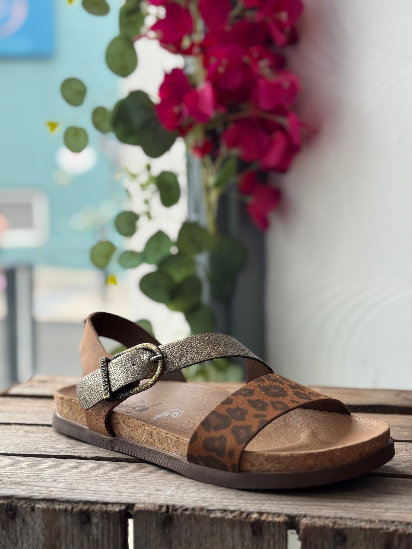 Brown sandal with leopard print pattern on a wooden surface with flowers in the background