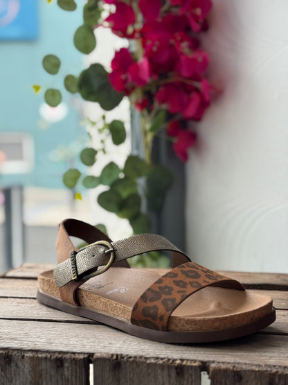 Brown sandal with leopard print pattern on a wooden surface with flowers in the background