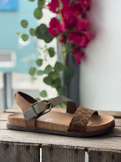 Brown sandal with leopard print strap on a wooden surface with flowers in the background