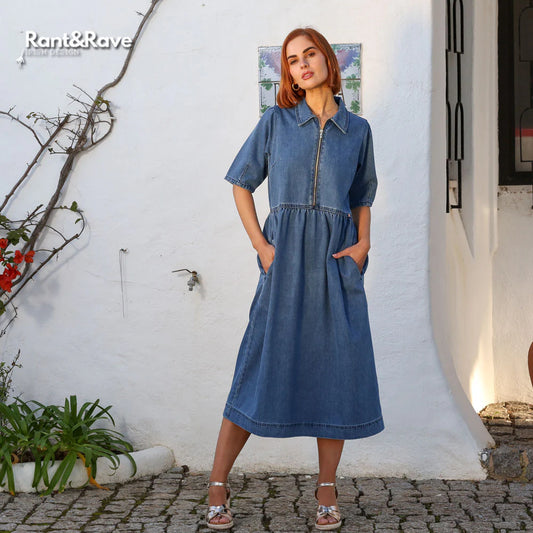 Woman wearing a denim dress standing against a white wall with plants around.