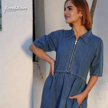 Woman wearing a blue denim dress standing against a white wall.