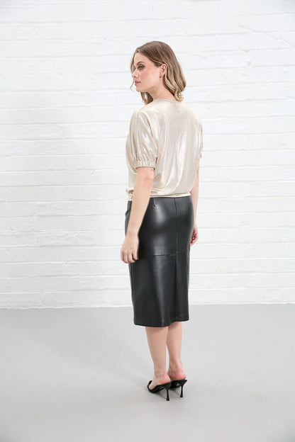 A woman stands in profile against a white brick wall, wearing a shiny champagne-colored short-sleeve top and a black faux-leather knee-length skirt with black high heels.