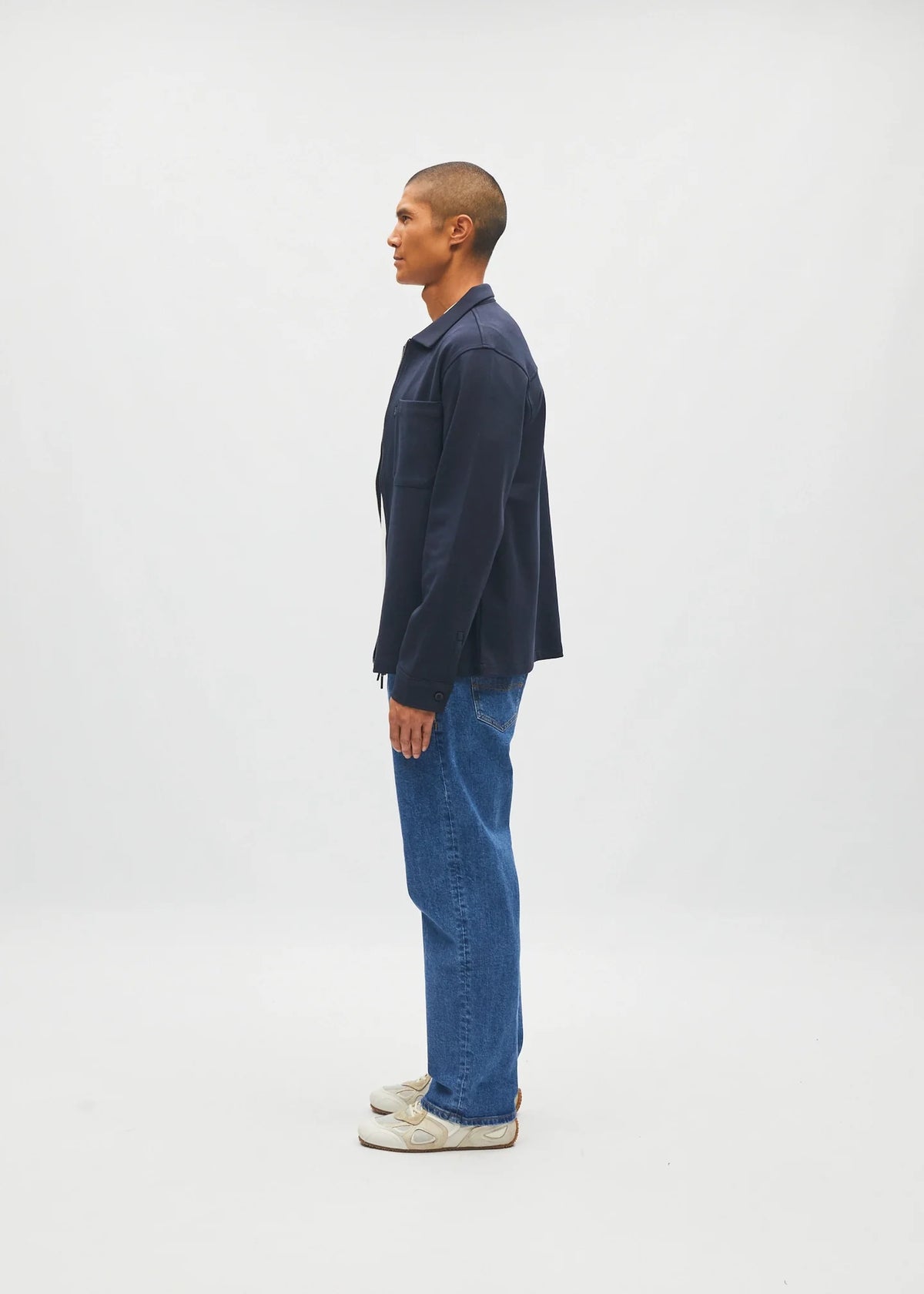 A man in profile wearing a dark navy blue overshirt, blue jeans, and white sneakers, standing against a plain light gray studio background.