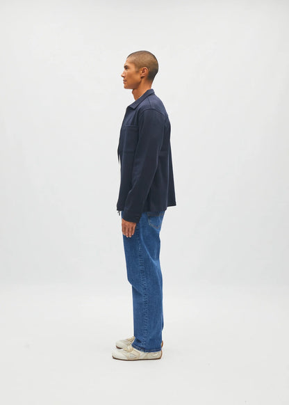 A man in profile wearing a dark navy blue overshirt, blue jeans, and white sneakers, standing against a plain light gray studio background.