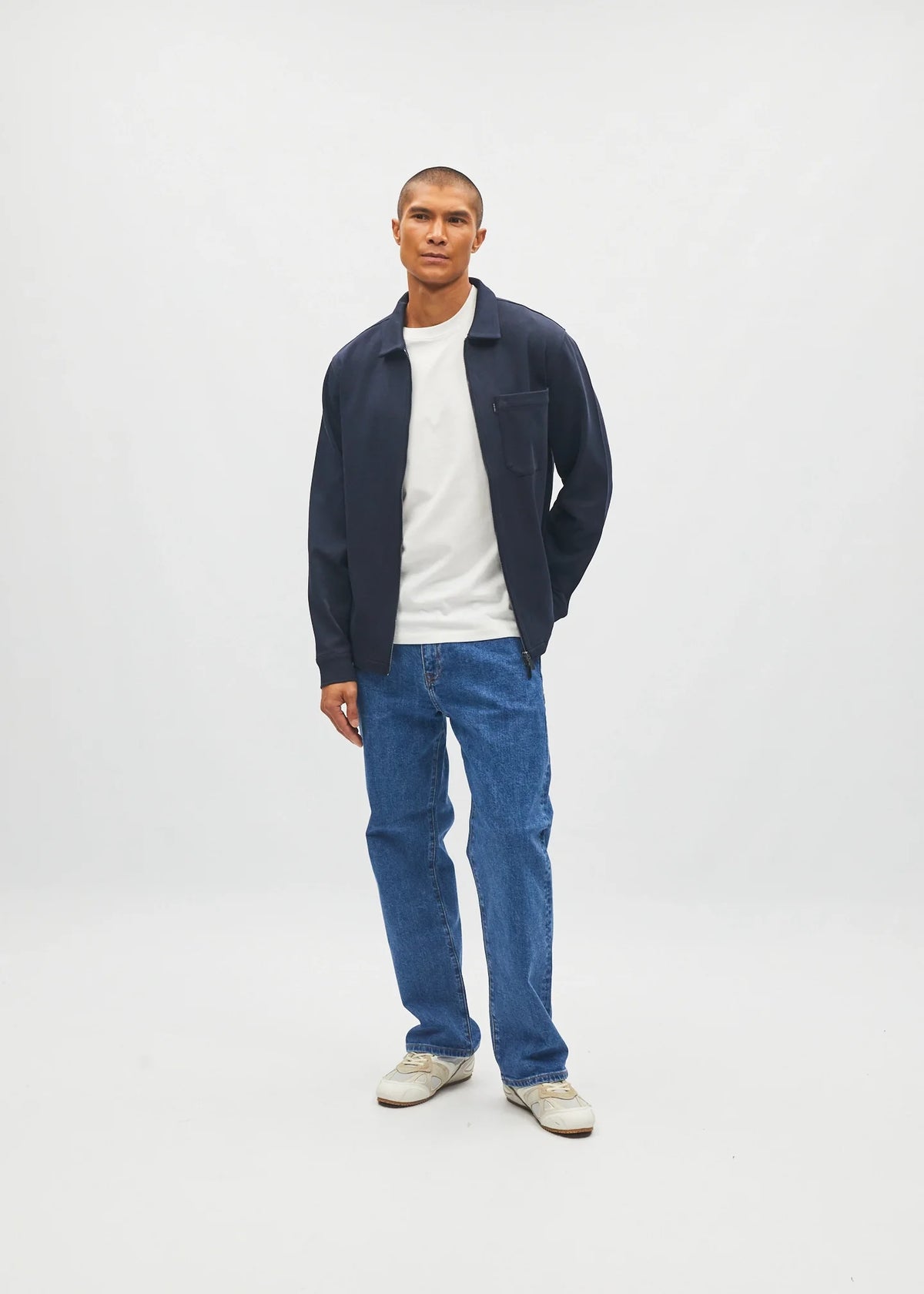 A man stands against a light gray studio background wearing a navy blue zip-up jacket over a white t-shirt, blue jeans, and off-white sneakers.