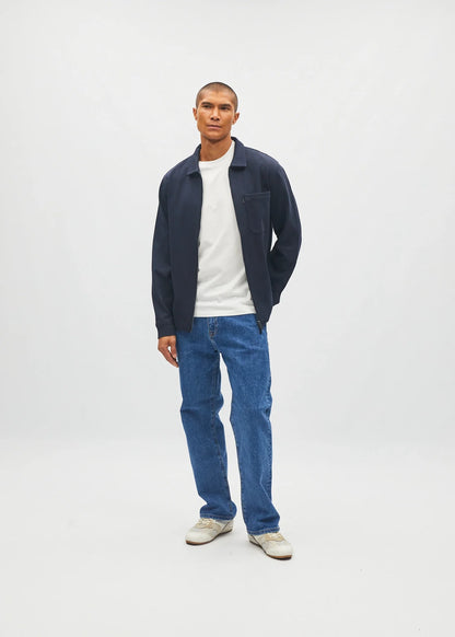 A man stands against a light gray studio background wearing a navy blue zip-up jacket over a white t-shirt, blue jeans, and off-white sneakers.