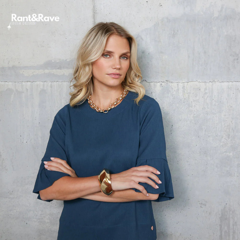 Woman wearing a blue top with gold jewelry against a gray concrete wall, featuring the brand 'Rant&Rave'.