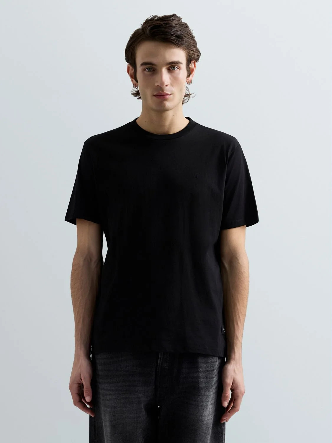 A man wearing a plain black short-sleeve crewneck t-shirt and dark gray jeans, standing against a light gray studio background.