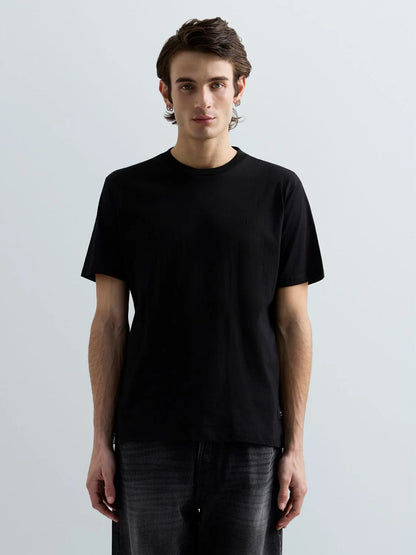 A man wearing a plain black short-sleeve crewneck t-shirt and dark gray jeans, standing against a light gray studio background.