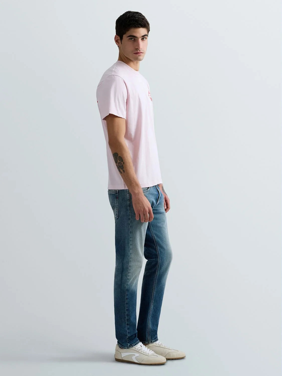 A man wearing a light pink crew-neck t-shirt, blue jeans, and off-white sneakers, standing in profile against a light gray studio background.
