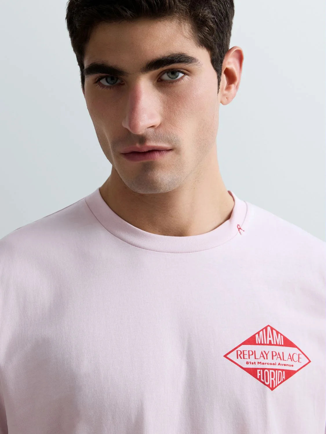 A close-up of a man wearing a light pink crewneck t-shirt with a red diamond-shaped 'Miami Replay Palace' graphic on the left chest, set against a light gray background.