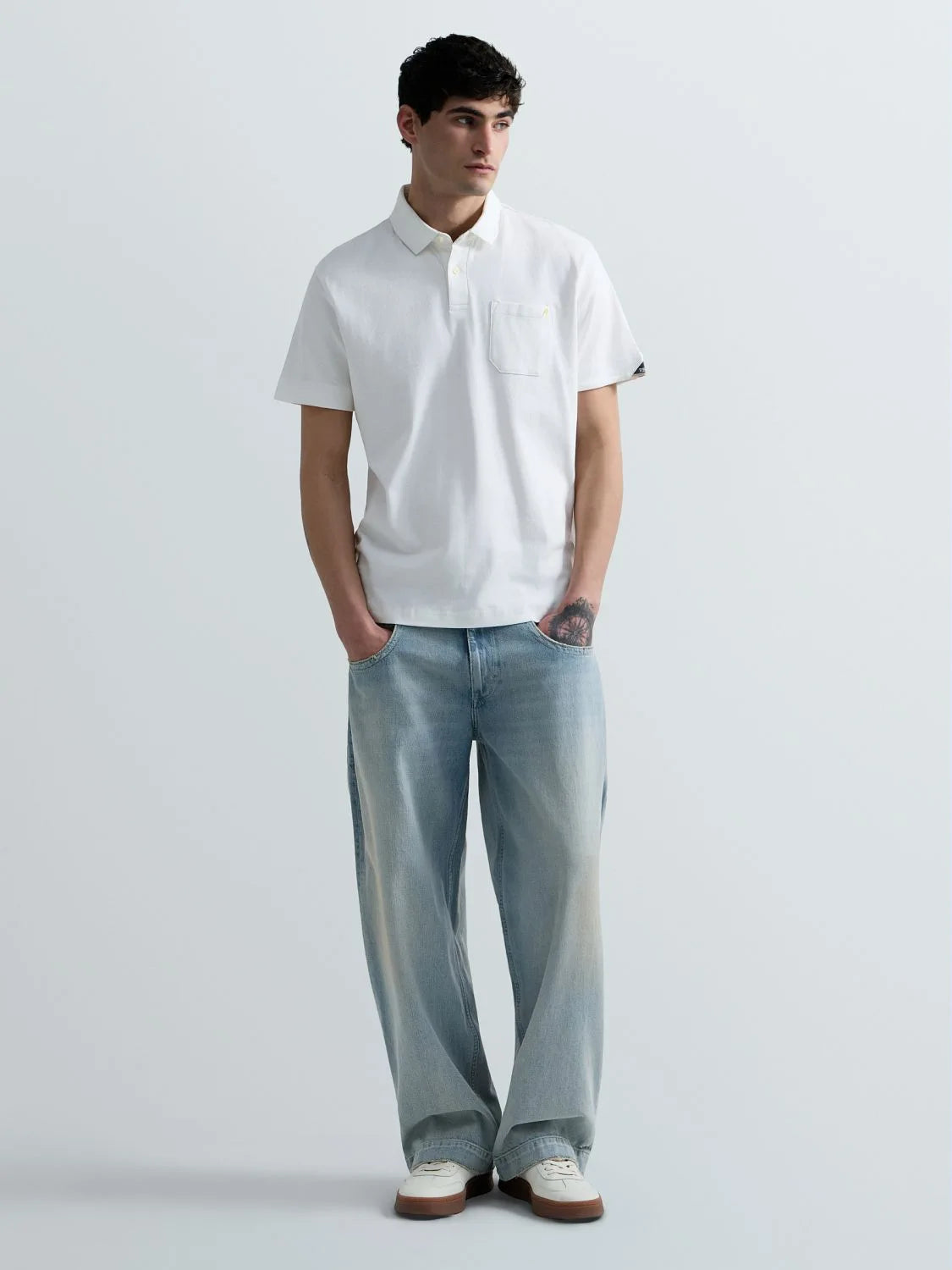 A man modeling a white short-sleeve polo shirt with a chest pocket, paired with light-wash wide-leg jeans and white sneakers, standing against a light gray studio background.