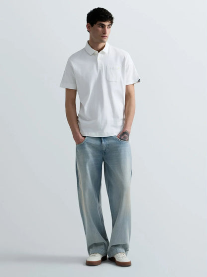 A man modeling a white short-sleeve polo shirt with a chest pocket, paired with light-wash wide-leg jeans and white sneakers, standing against a light gray studio background.