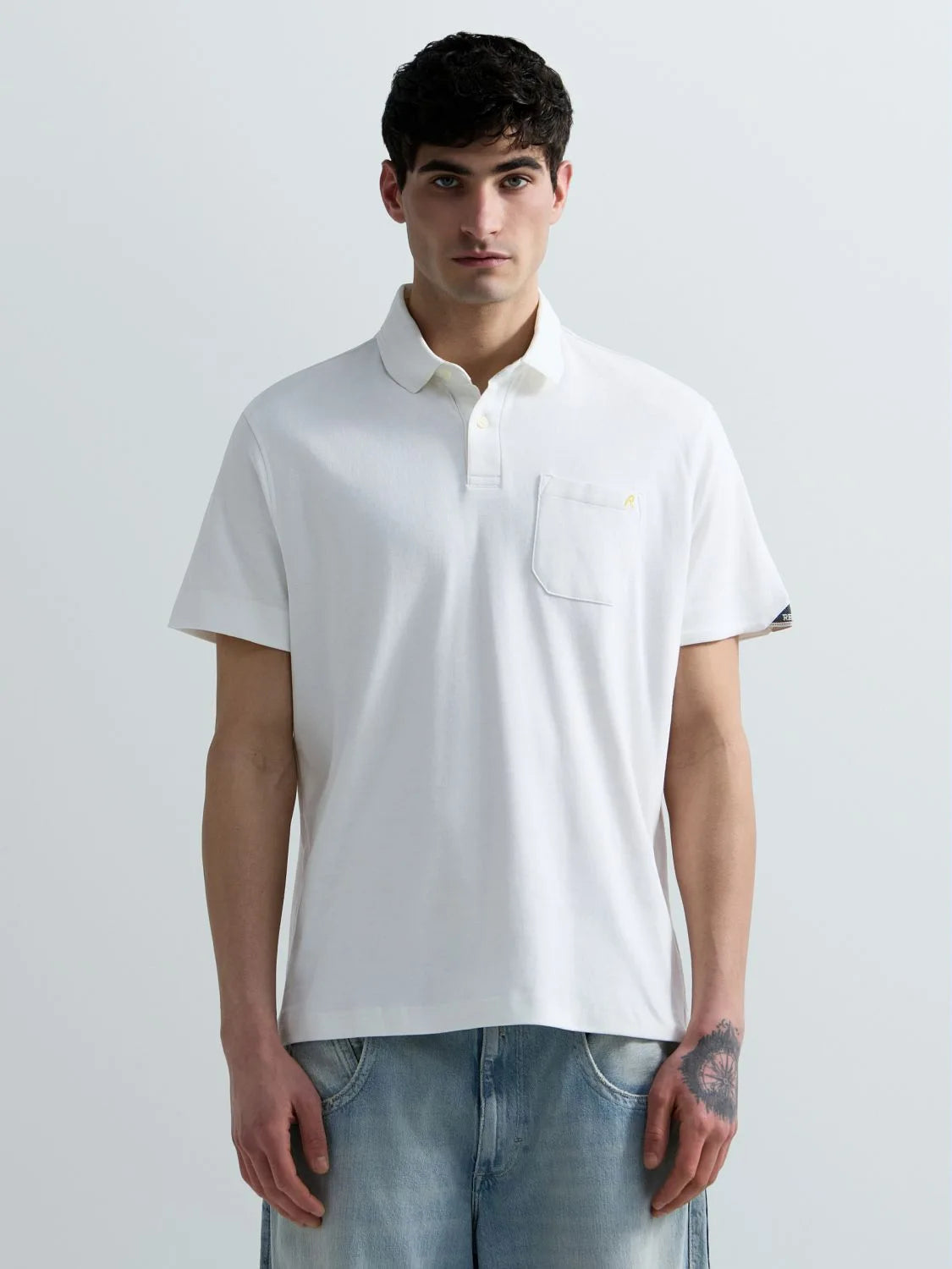 A man wearing a white short-sleeve polo shirt with a chest pocket and light blue jeans, standing against a light gray studio background.