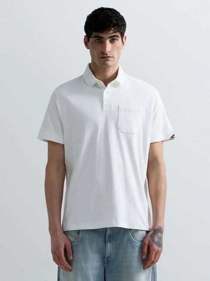 A man wearing a white short-sleeve polo shirt with a chest pocket and light blue jeans, standing against a light gray studio background.