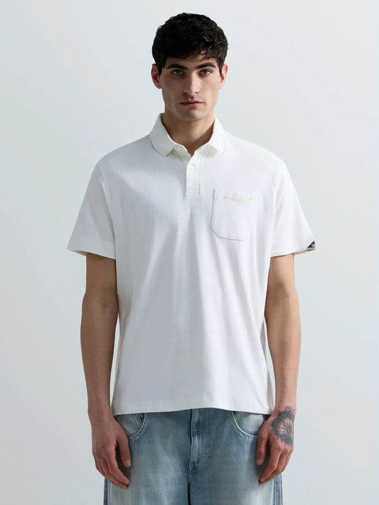 A man wearing a white short-sleeve polo shirt with a chest pocket and light blue jeans, standing against a light gray studio background.