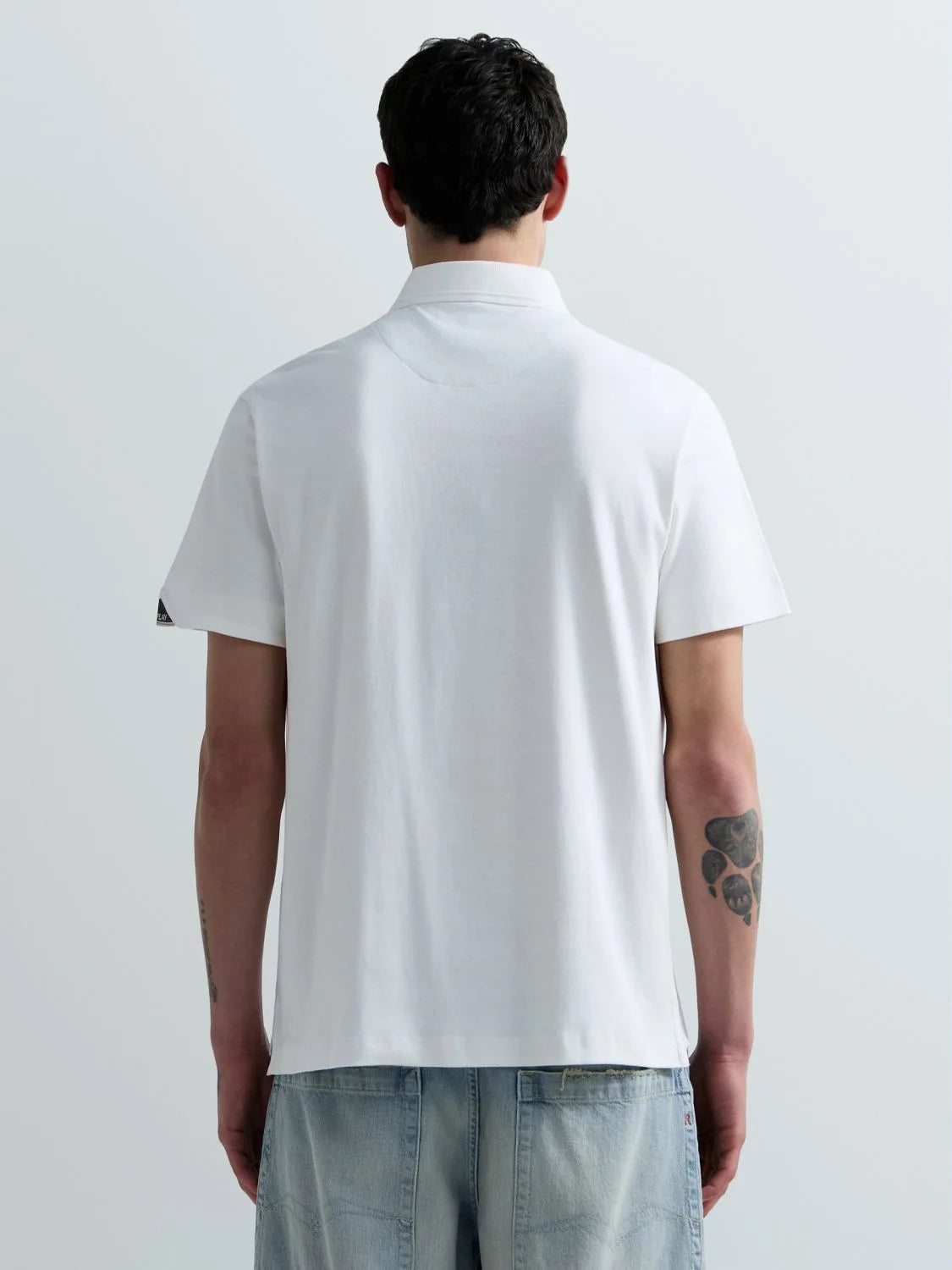A man viewed from behind wearing a white short-sleeve polo shirt and light blue jeans against a plain light gray studio background.