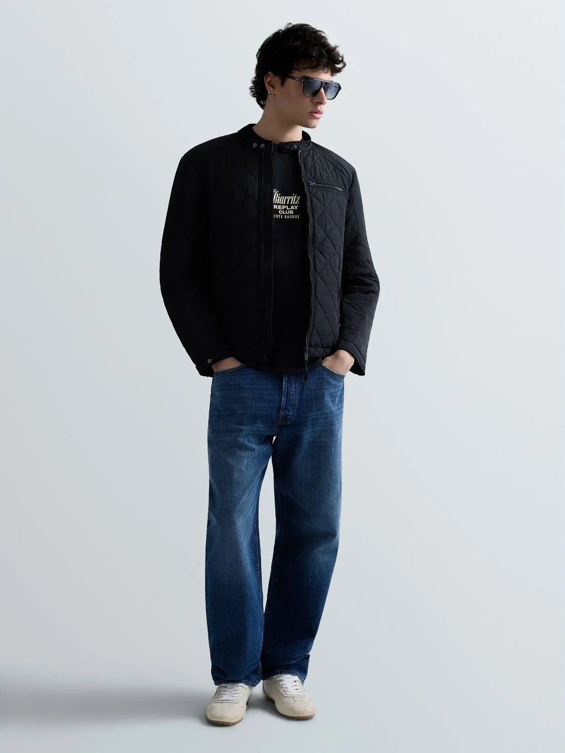 A man wearing a black quilted jacket over a black t-shirt, blue jeans, and white sneakers, standing against a light gray studio background.