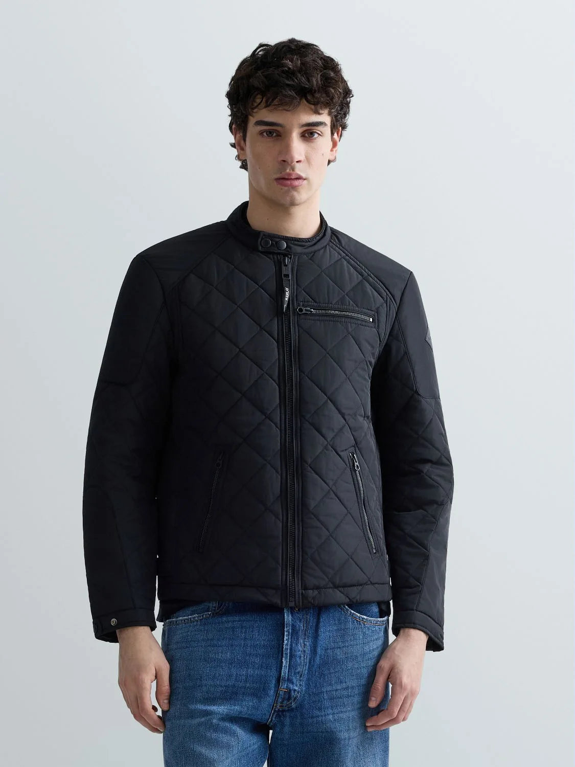 A man wearing a black diamond-quilted jacket with zippered pockets, paired with blue jeans, standing against a light gray studio background.