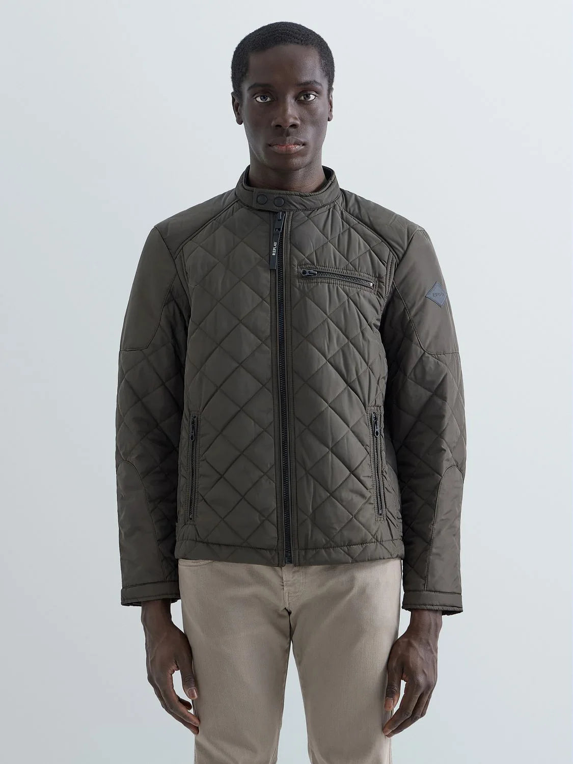 A man wearing a dark olive green diamond-quilted jacket with a high collar and front zipper, paired with beige trousers, standing against a light gray studio background.