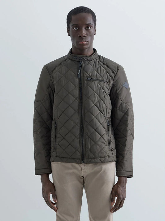A man wearing a dark olive green diamond-quilted jacket with a high collar and front zipper, paired with beige trousers, standing against a light gray studio background.