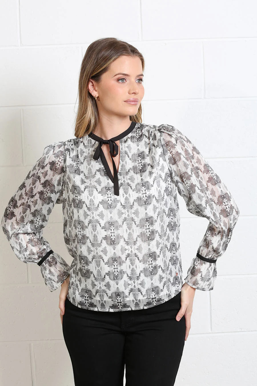 A woman wearing a sheer long-sleeve blouse with a gray, black, and white abstract pattern, paired with black pants, standing against a white brick wall.