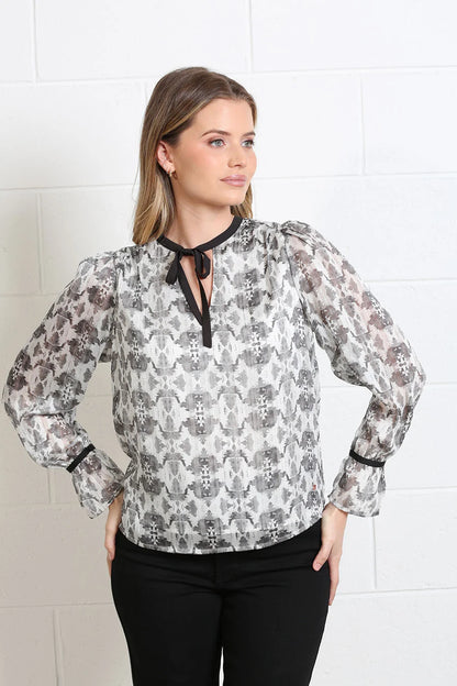 A woman wearing a sheer long-sleeve blouse with a gray, black, and white abstract pattern, paired with black pants, standing against a white brick wall.