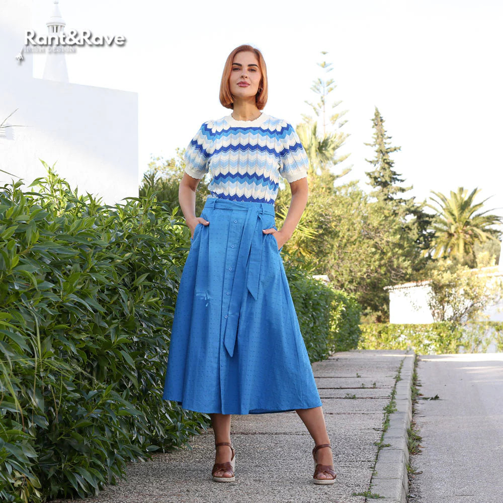 Woman wearing a blue and white striped shirt and blue skirt standing on a path with greenery.