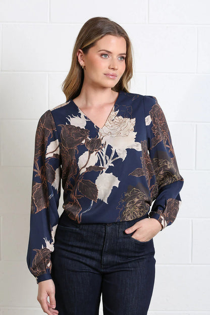 A woman wearing a navy blue long-sleeve blouse with a beige and brown floral print, paired with dark blue jeans, standing against a white brick wall.
