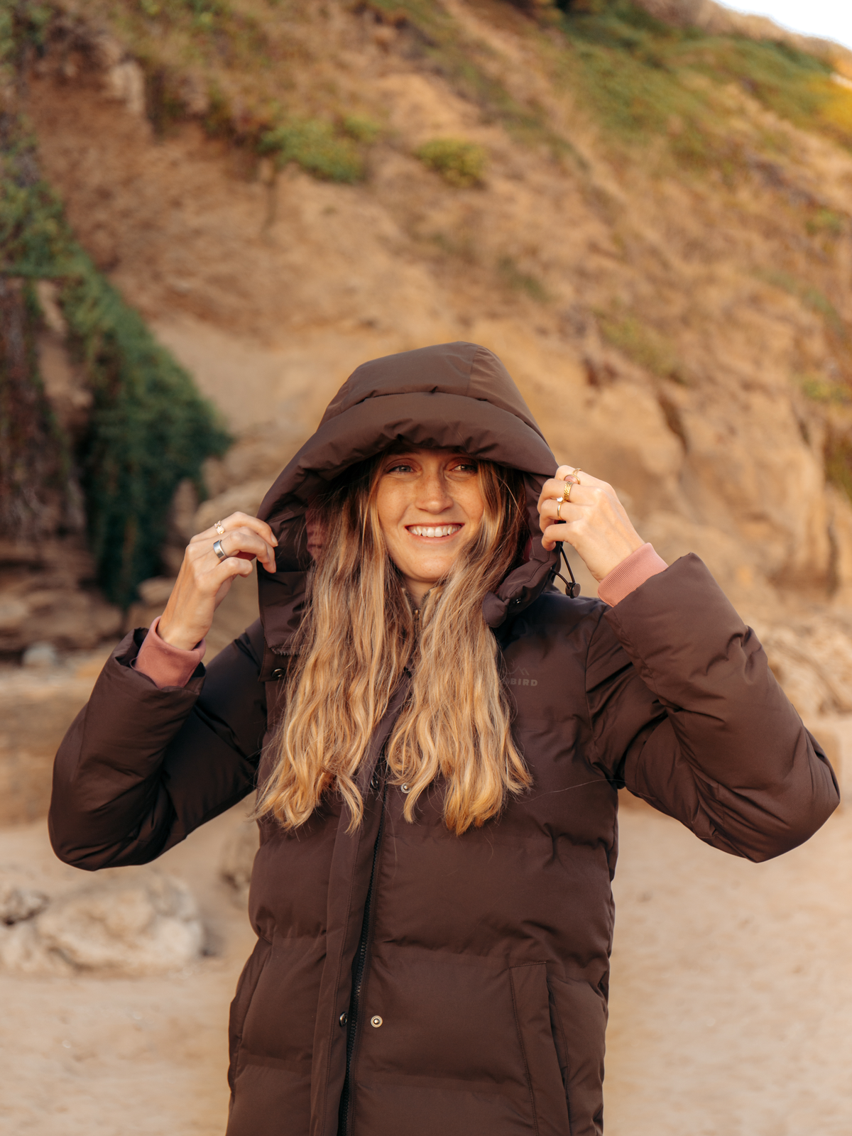 A woman wearing a dark brown hooded puffer jacket stands outdoors on a sandy path with a rocky hill in the background, smiling and adjusting the hood.