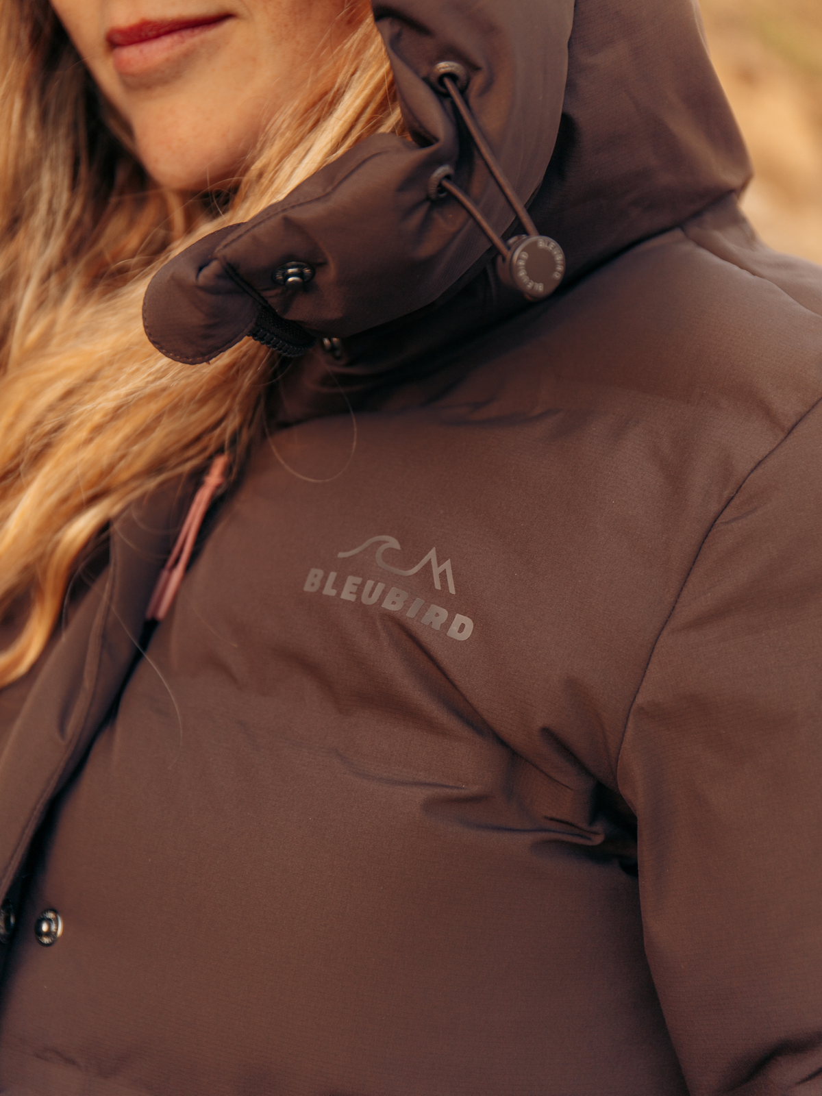 Close-up of a person wearing a dark brown hooded puffer jacket with a visible Bleubard logo on the chest, photographed outdoors with warm natural light.