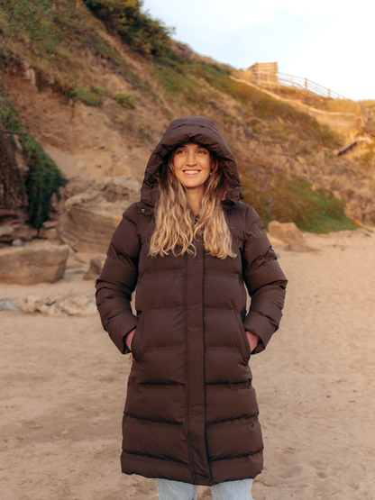 A woman wearing a long, dark brown hooded puffer coat stands on a sandy beach with a rocky hillside in the background.