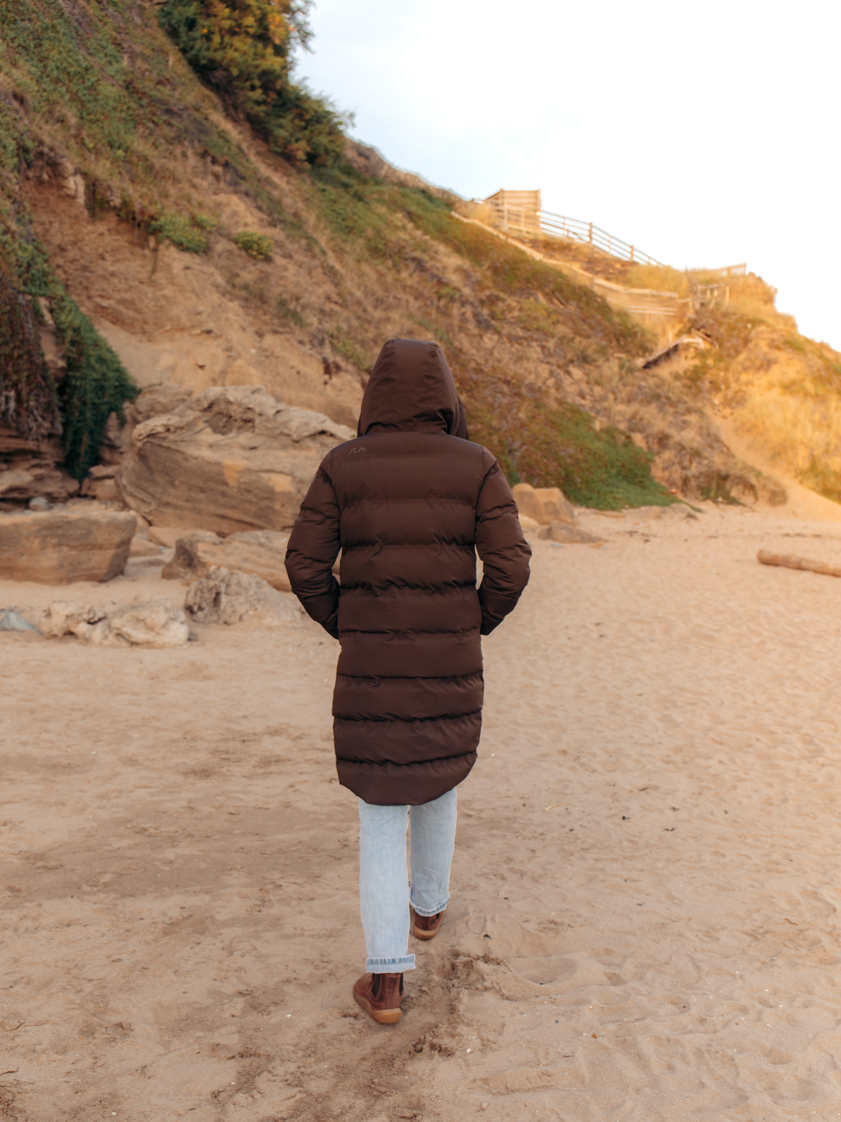 A person viewed from behind wearing a long brown puffer coat, light blue jeans, and brown shoes, walking on a sandy beach with a rocky, grassy cliff in the background.