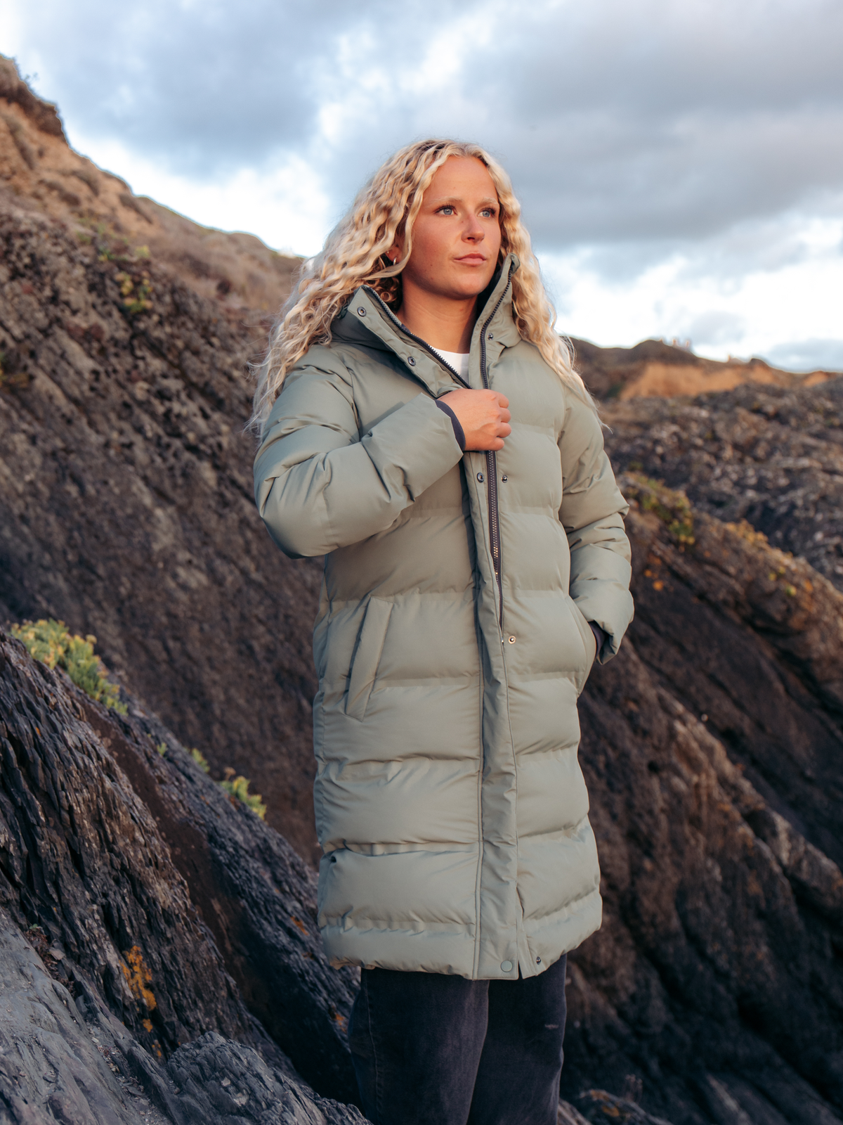 A woman wearing a long, light sage green puffer coat stands outdoors on a rocky mountain slope under a cloudy sky.