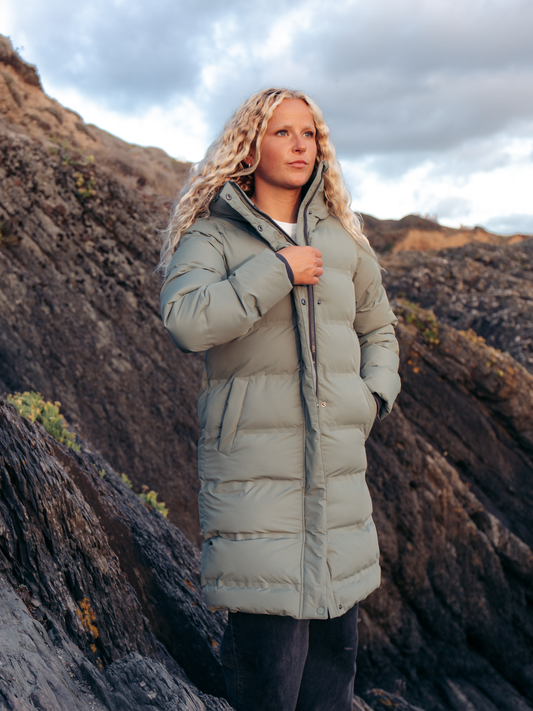 A woman wearing a long, light sage green puffer coat stands outdoors on a rocky mountain slope under a cloudy sky.