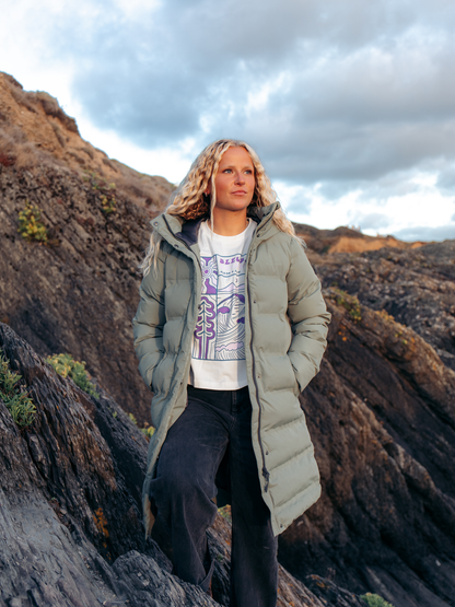 A woman stands on a rocky mountain slope wearing a long light sage green puffer coat over a white graphic t-shirt and dark pants, with a dramatic cloudy sky behind her.