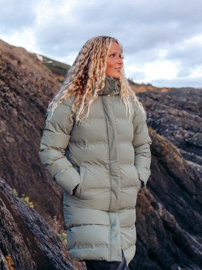 A woman with long curly blonde hair stands outdoors wearing a long, quilted puffer coat in a muted sage green color, set against a rocky, dark volcanic landscape under a cloudy sky.