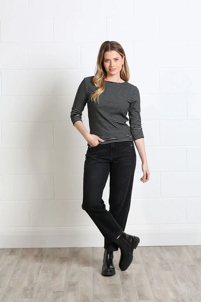 A woman modeling a black and white striped long-sleeve top with black jeans and black boots, standing against a white brick wall and light wood floor.