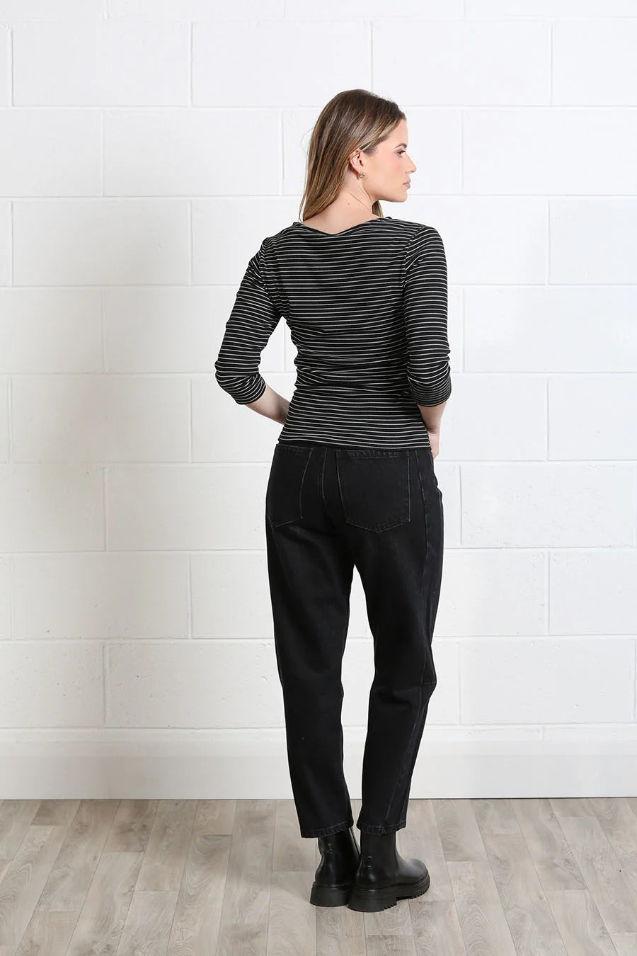 A woman is shown from the back wearing a black and gray striped long-sleeve top, black jeans, and black boots, standing against a white brick wall.