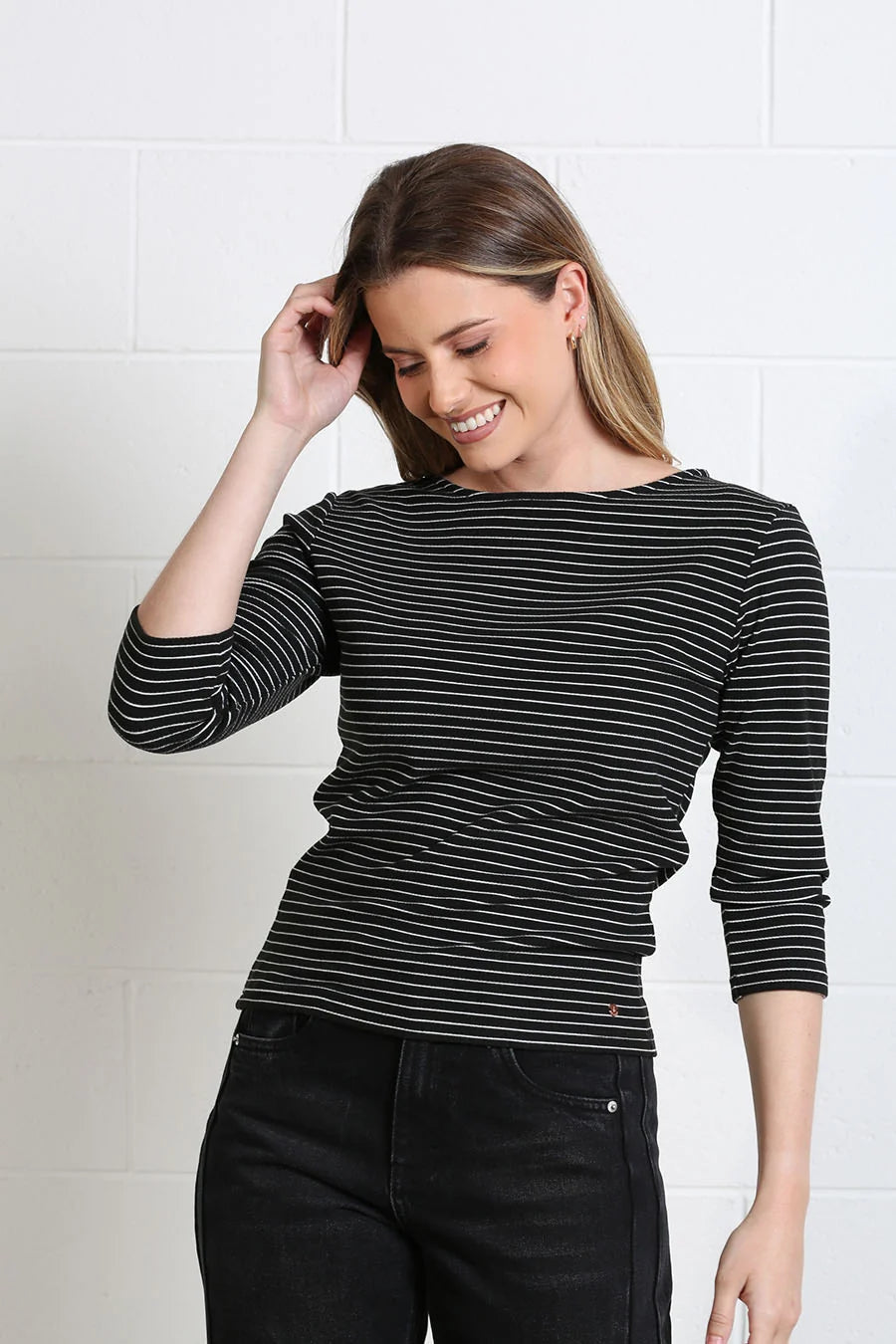 A woman wearing a black and white horizontally striped long-sleeve top and black jeans, standing against a white brick wall.