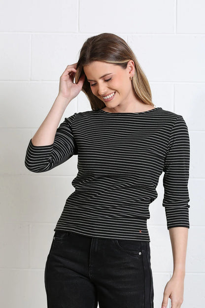 A woman wearing a black and white horizontally striped long-sleeve top and black jeans, standing against a white brick wall.