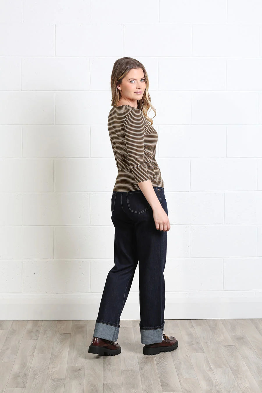 A woman stands in profile against a white brick wall and light wood floor, wearing a brown and beige striped three-quarter sleeve top, dark blue jeans with rolled cuffs, and dark brown shoes.