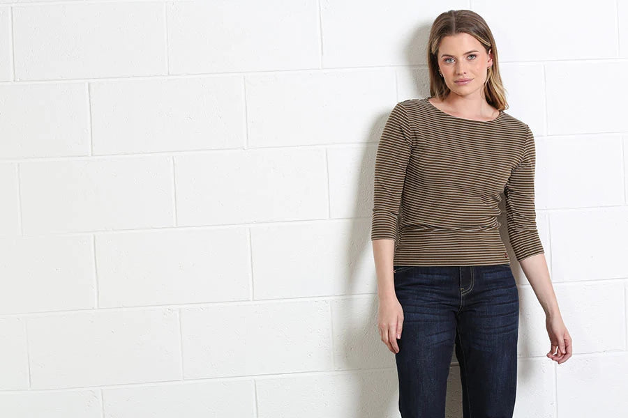 A woman wearing a brown and beige horizontally striped three-quarter sleeve top and dark blue jeans, standing against a white painted brick wall.