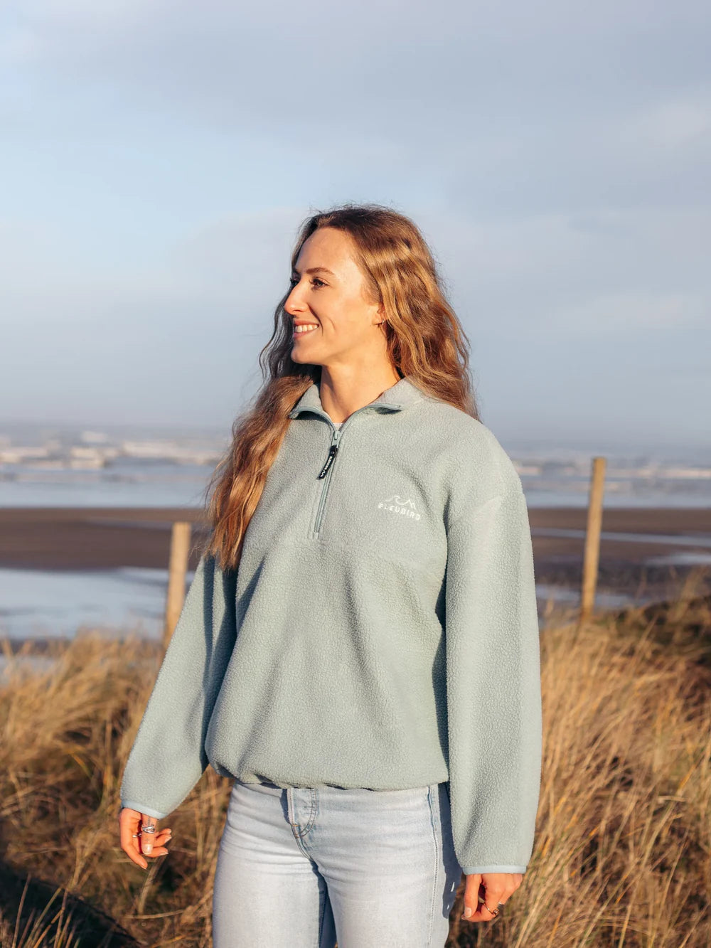 Woman wearing a light gray fleece jacket with a logo, standing in a field with a scenic background.