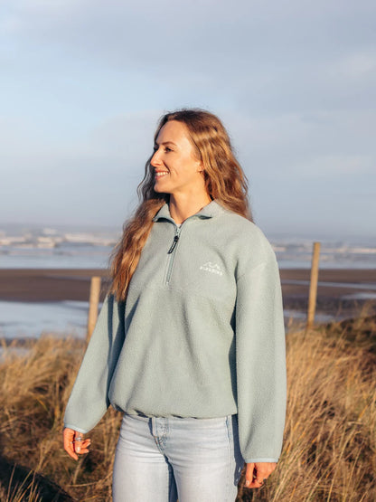 Woman wearing a light gray fleece jacket with a logo, standing in a field with a scenic background.