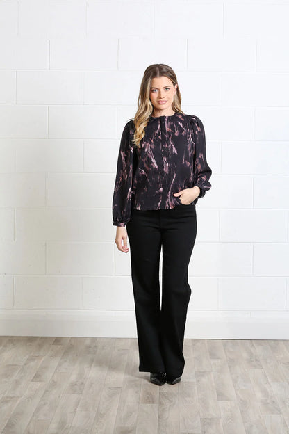 A woman modeling a long-sleeve black blouse with a dusty pink abstract marble pattern, paired with black wide-leg pants, standing against a white brick wall and light wood floor.